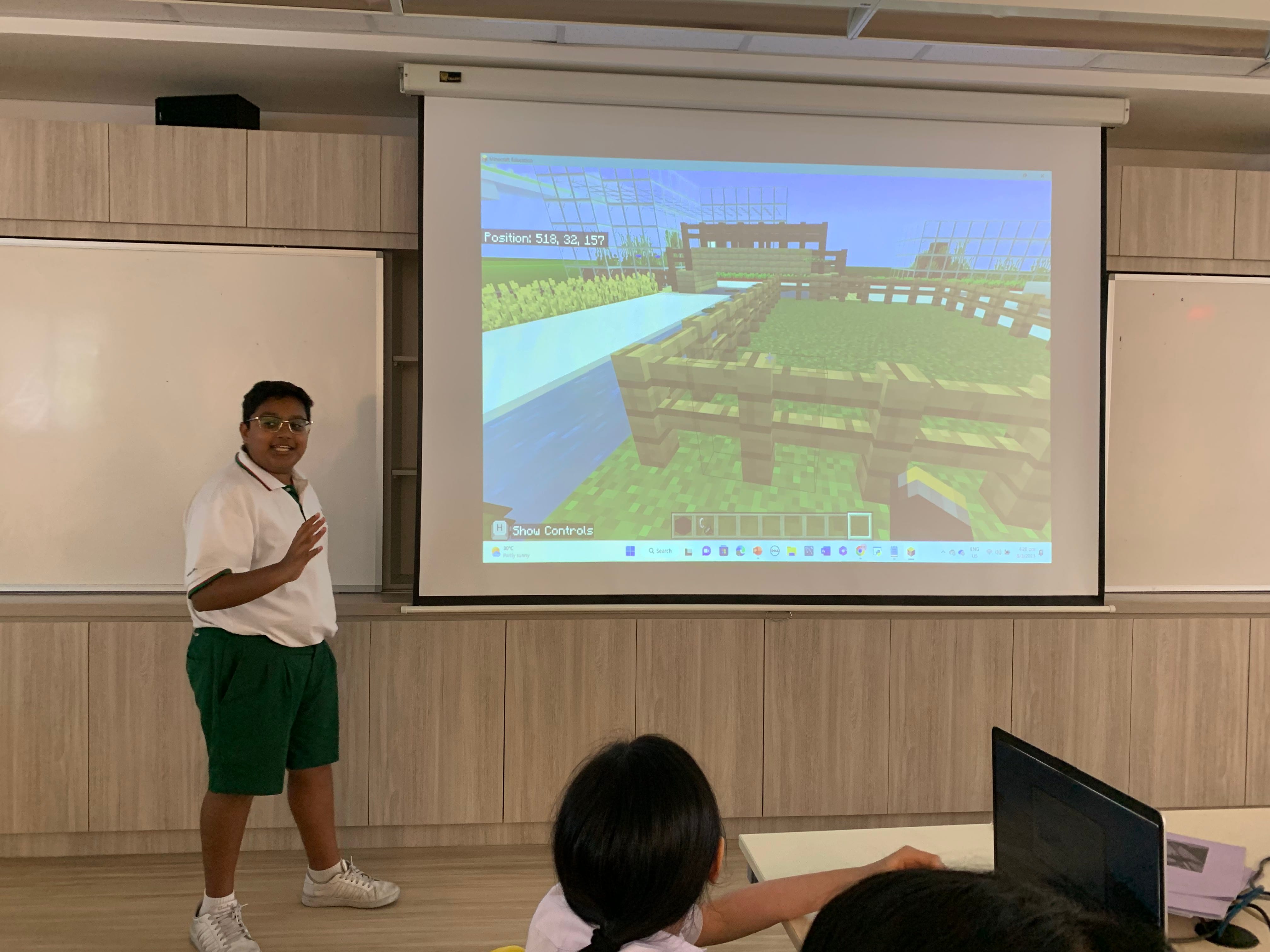 A student in a white shirt and green shorts presents a Minecraft game screenshot on a classroom projector screen.